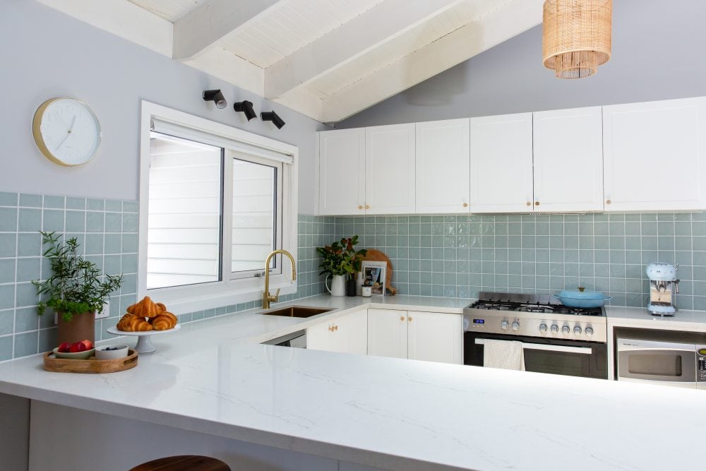 White shaker style kitchen cabinetry with brass knob handles and stone benchtop and acqua tiles splashback