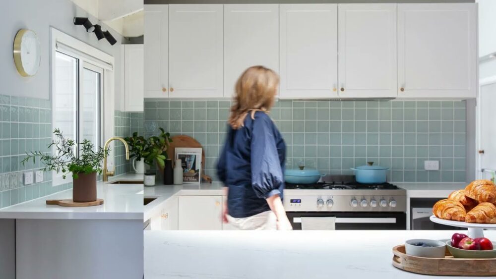 Lady dressed in blue shirt walking through a u shaped white kitchen with green tiled splashback with fruit and pastries on bench