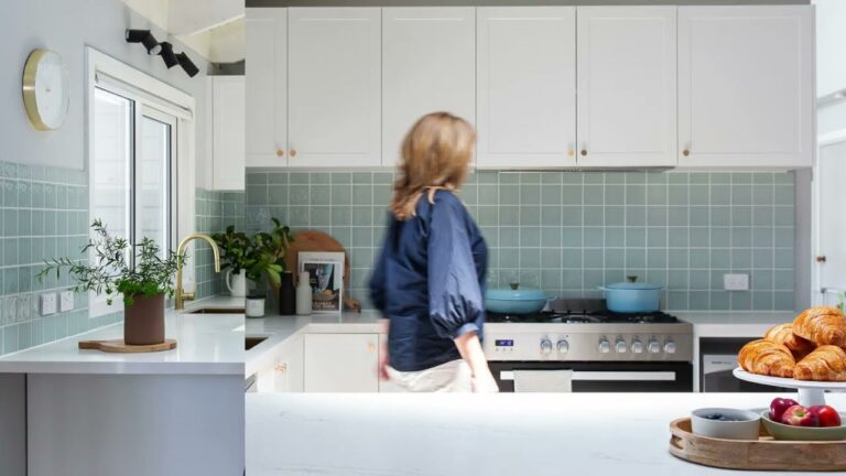 Lady dressed in blue shirt walking through a u shaped white kitchen with green tiled splashback with fruit and pastries on bench