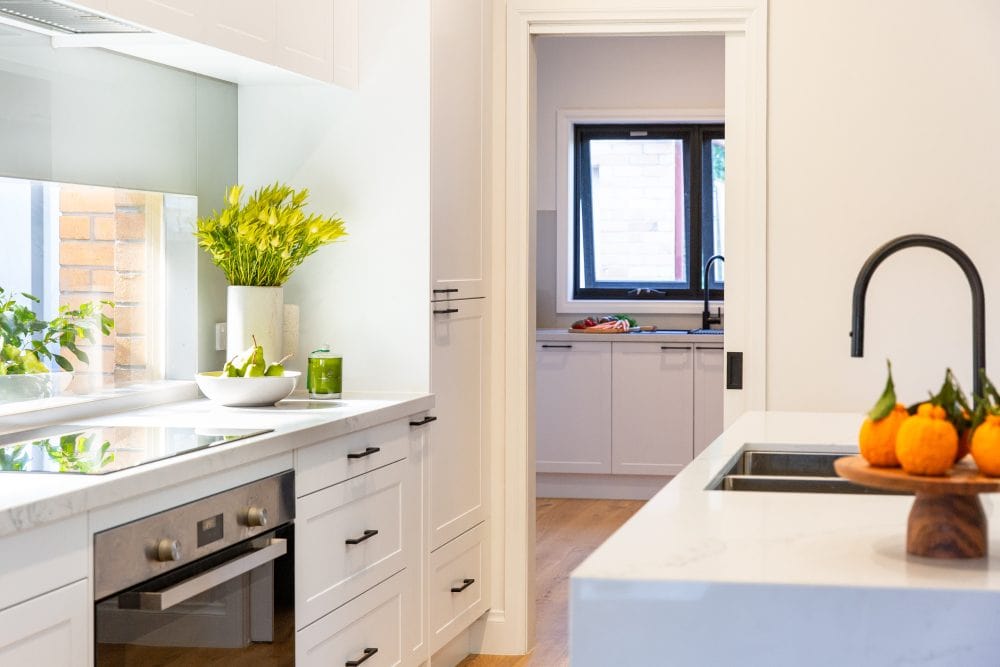 Galley kitchen with white shaker style cabinetry and large white island bench with black sink and tap and a timber stand with tangelos on the benchtop