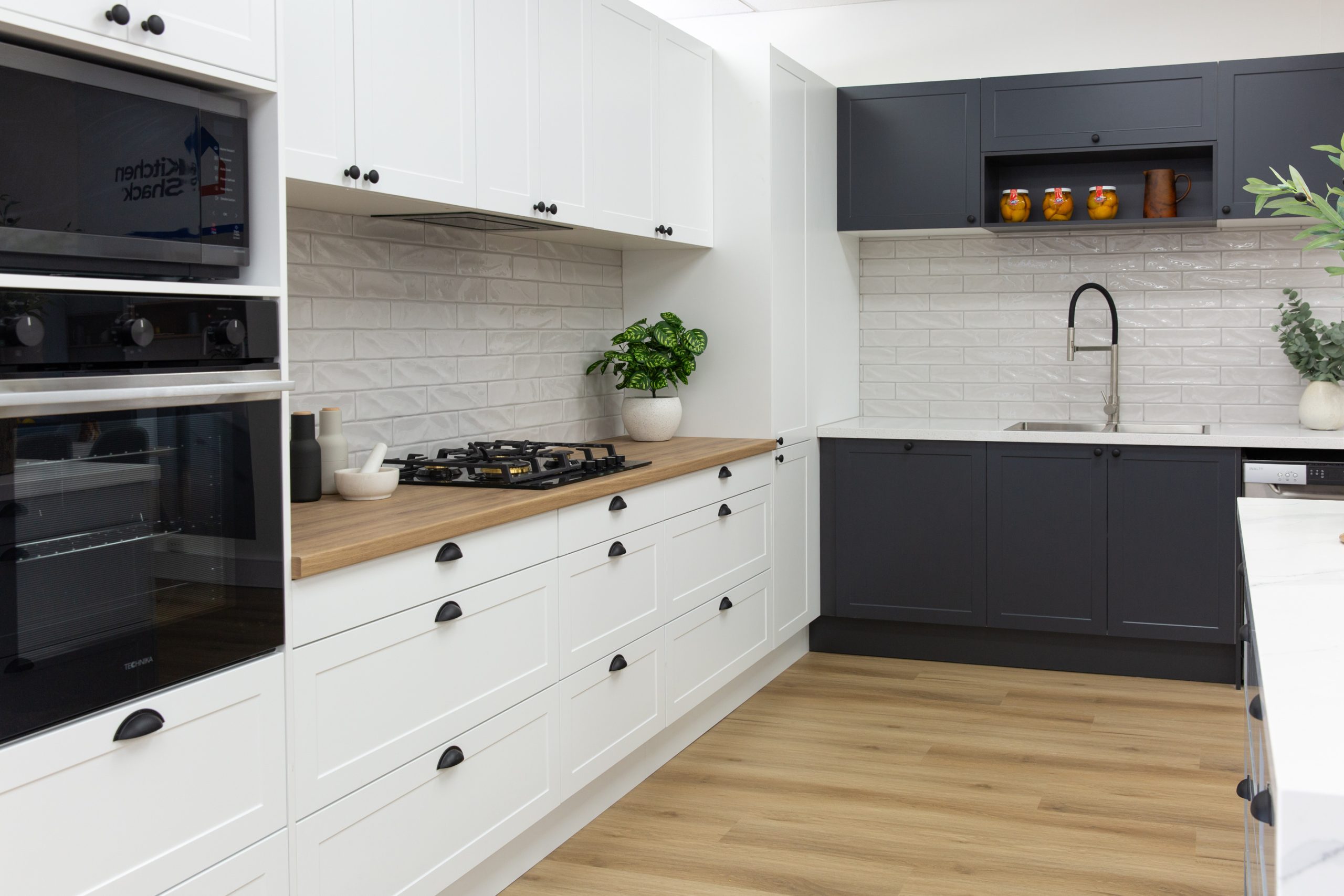 Showroom display featuring shaker style white and black kitchen cabinetry with black ovens and timber benchtops