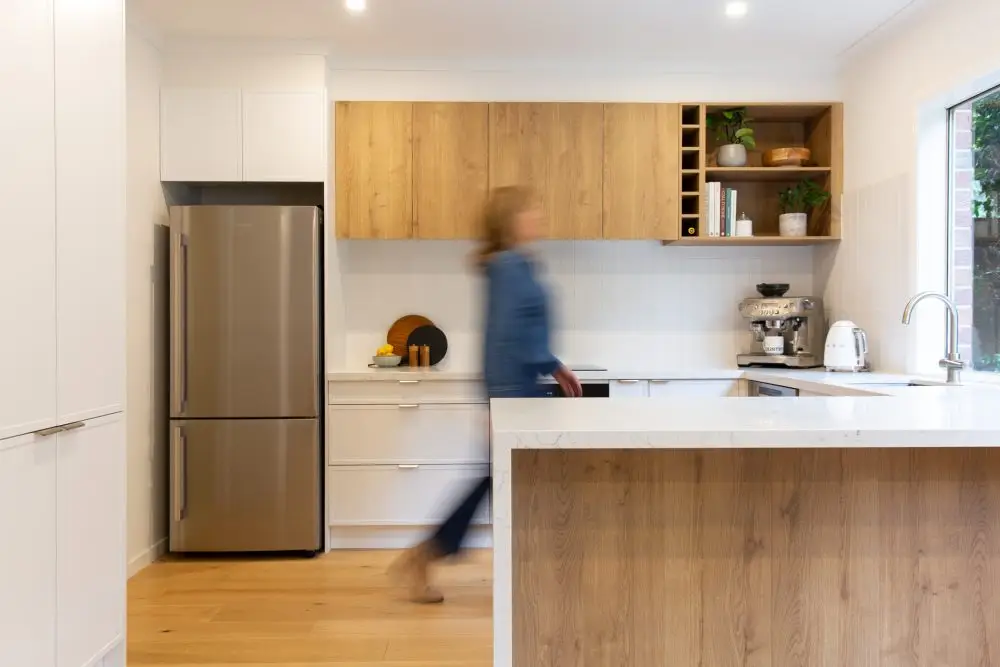 Female walking through galley kitchen with white kitchen cabinetry and oak timber look overhead cabinetry and island bench with white stone benchtops
