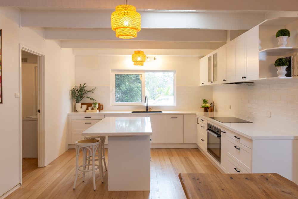 Modern white kitchen with shaker style cabinetry, timber flooring and central island bencht with stools. Features black oven and cooktop, open shelves with plants and two rattan pendant lights