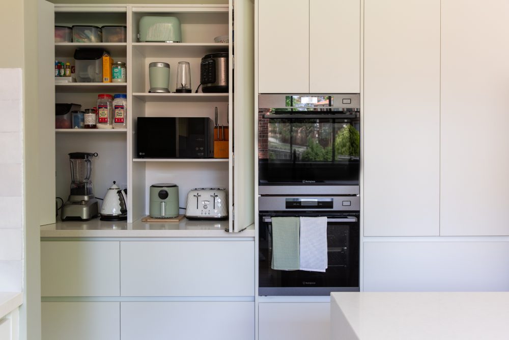 Double appliance cabinet containing small appliances and pantry staples in a white handle free kitchen next to double ovens