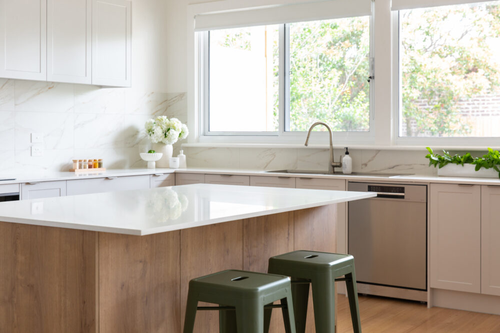 Kitchen displaying large island bench in the centre with overhang in white stone and underneath oak timber cabinetry with two green benchstools
