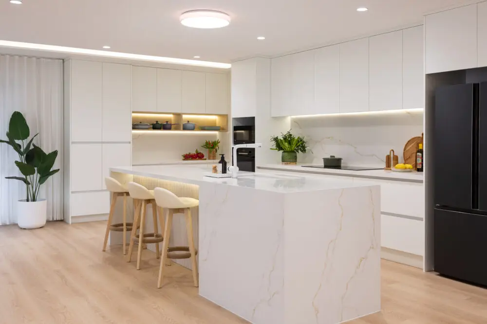 Large open plan kitchen featuring white cabinetry and standalone stone island bench with black oven and microwave and vase of flowers and pots decorating benchtops and open shelf