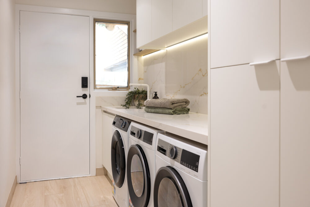 Galley laundry with white cabinetry and side by side washer dryers with green towels and hand soap on stone benchtop