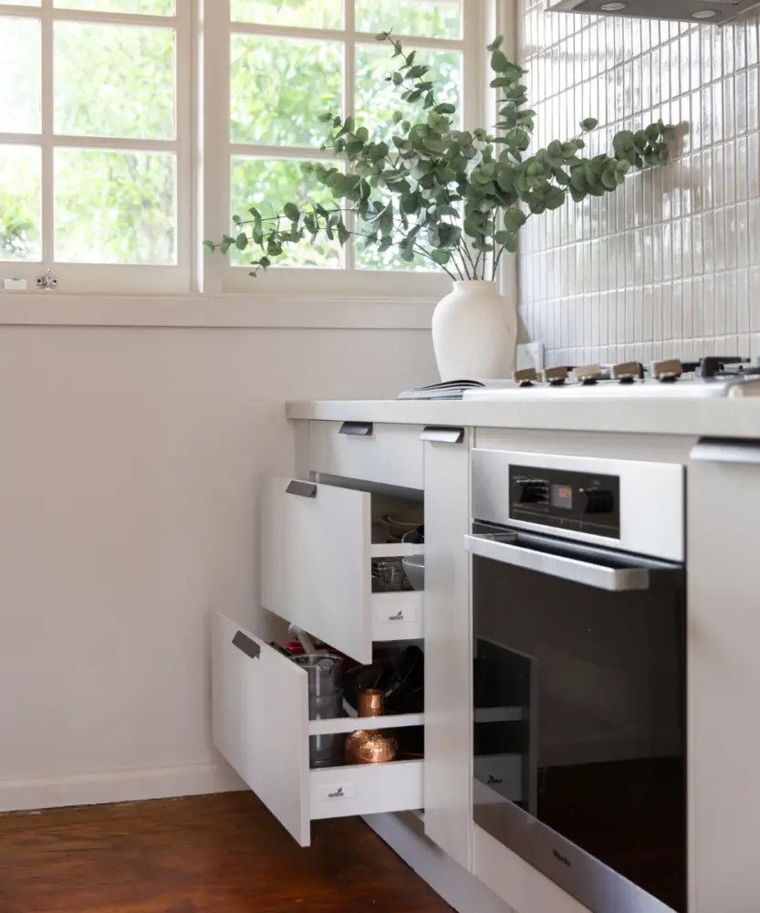 White kitchen displaying 3 drawers open with stone benchtops and vase with folliage on bench