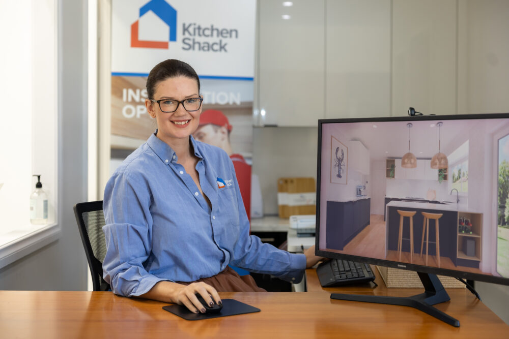 Lady in a blue shirt sitting at a desk with the computer designing a kitchen space in blue. A Kitchen Shack logo sign in the background.