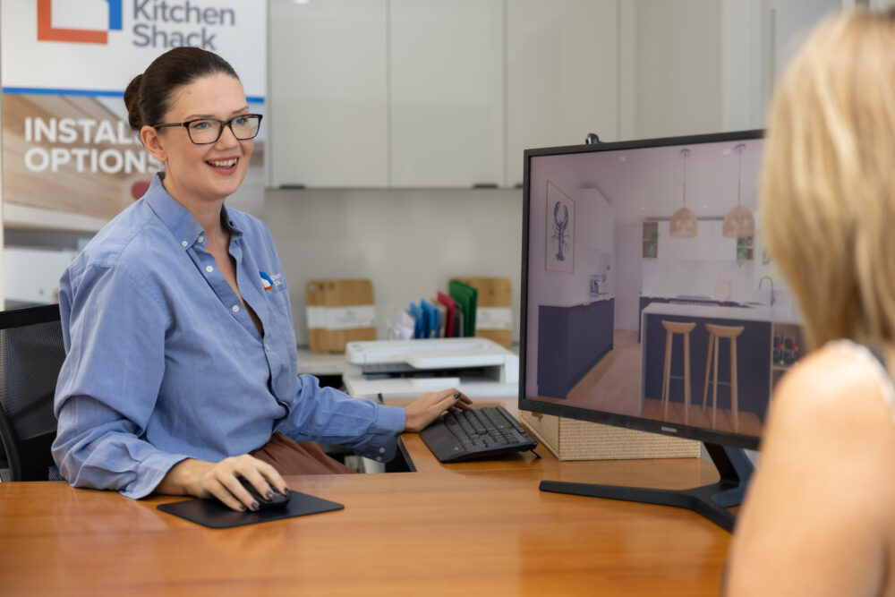 Female dressed in a blue shirt sitting at desk designing a kitchen in blue and timber with customer viewing