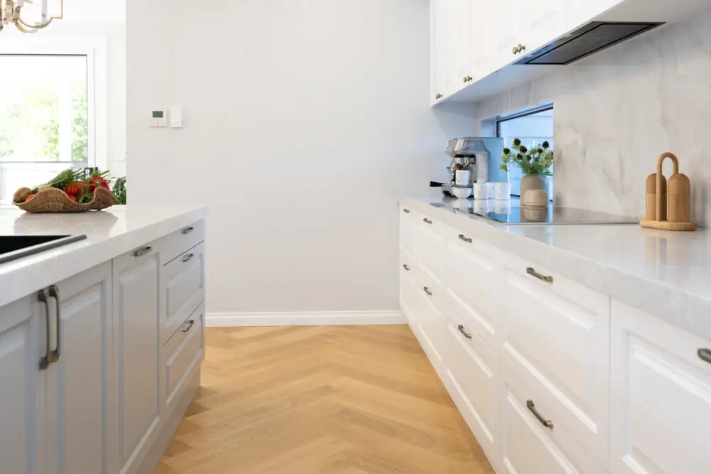 Galley style kitchen featuring white and grey kitchen cabinetry in carrington style with chrome handles and coffee machine with cups on benchtops as well as vase of flowers and styling items