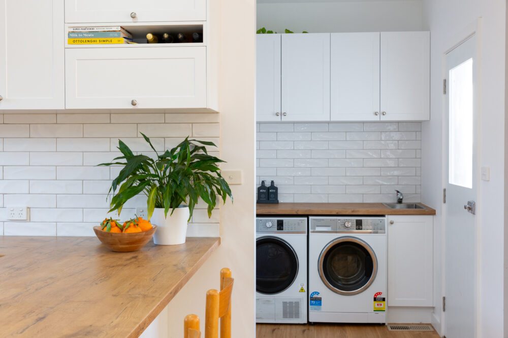Kitchen and laundry next to each other in white cabinetry with timber laminate oak benchtops featuring side by side washer dryer and timber bar stool with plant and bowl of oranges on display on benchtop