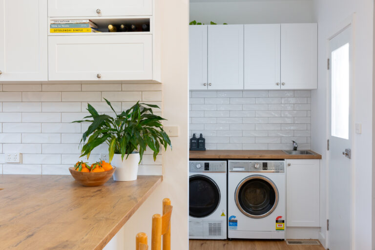 Kitchen and laundry next to each other in white cabinetry with timber laminate oak benchtops featuring side by side washer dryer and timber bar stool with plant and bowl of oranges on display on benchtop