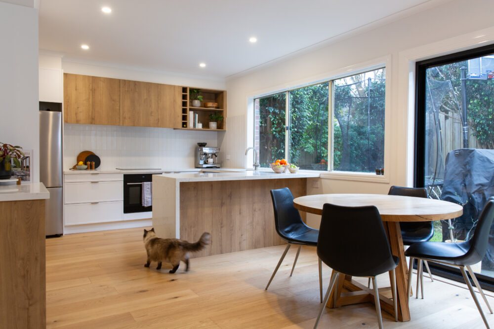 Galley kitchen displaying white hampton door and urban oak overhead cabinetry and timber in front of island bench with timber round dining table with black chairs and brown and white cat wandering through