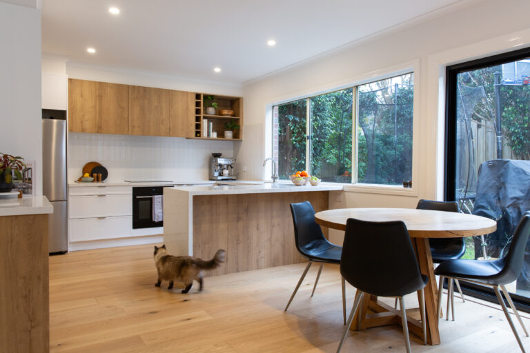 Galley kitchen displaying white hampton door and urban oak overhead cabinetry and timber in front of island bench with timber round dining table with black chairs and brown and white cat wandering through