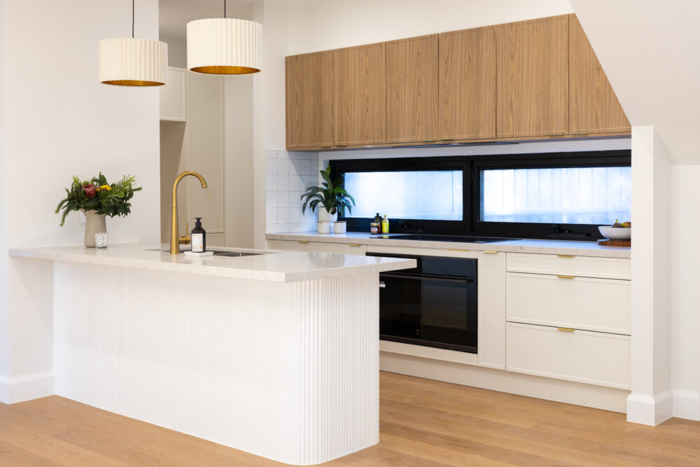 White and timber galley kitchen featuring black windows and oven with two pendant lights with brass tapware and flowers on the bench for styling