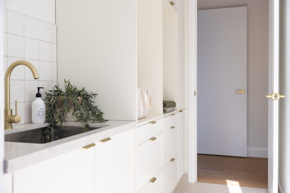 Galley style laundry with white cabinetry and brass handles with a plant on bench next to sink and tap
