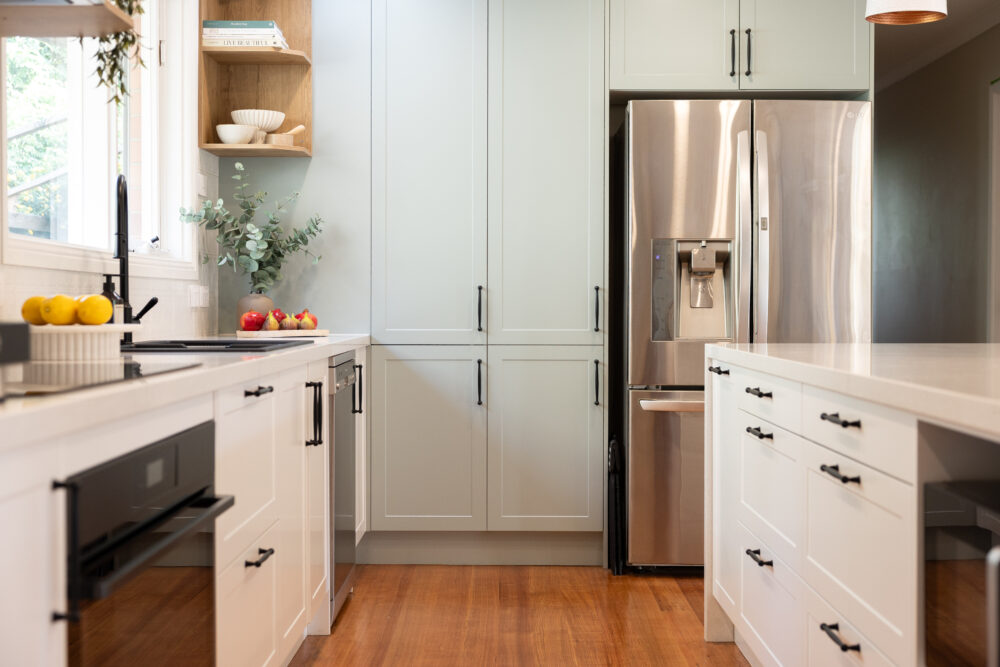 White and green galley kitchen with black handles and end pantry next to an open shelf displaying cookbooks and fruit and folliage on bench