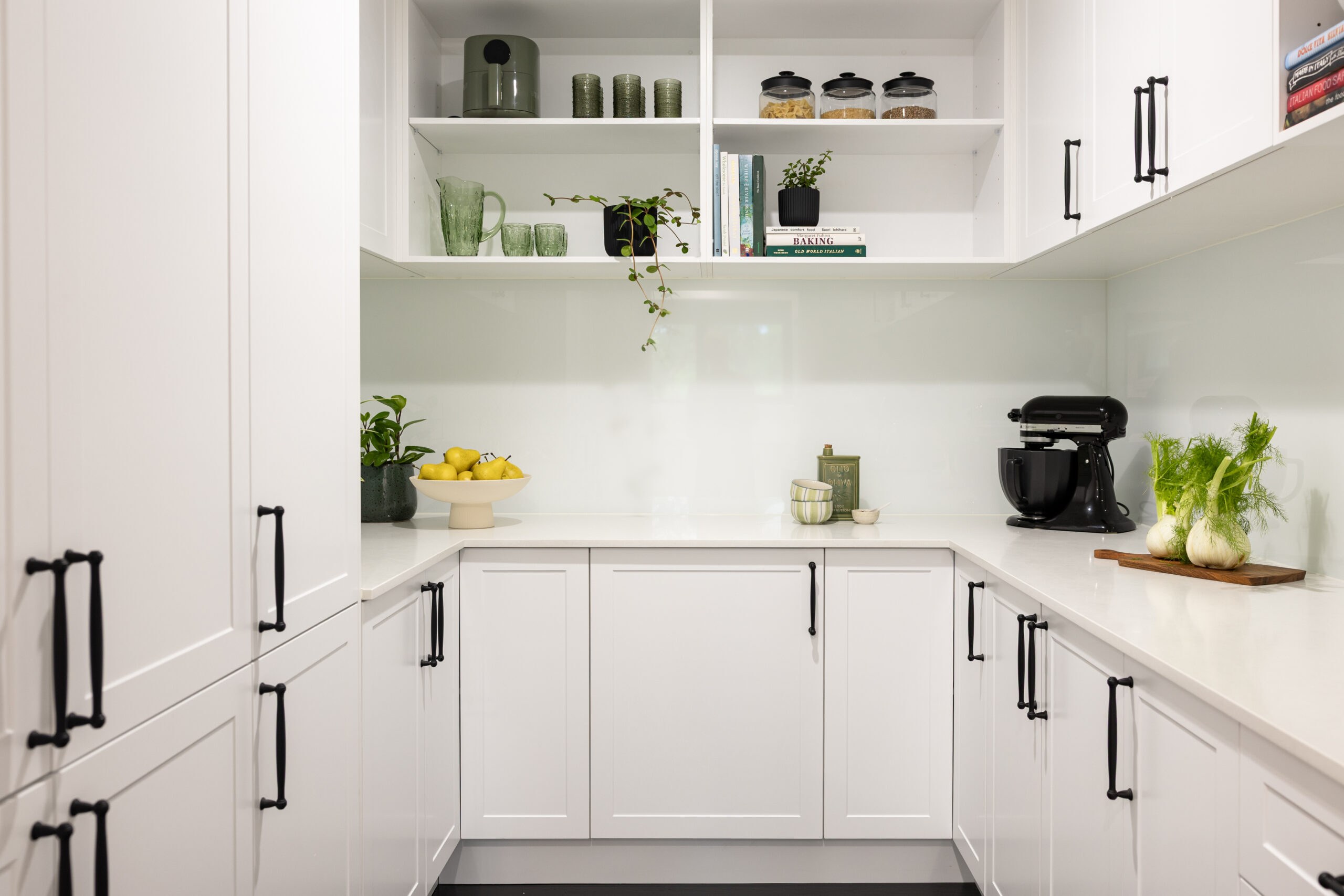 U shaped butlers pantry with white shaker style cabinetry with black handles and open shelves displaying green glasses, plant and styling items on stone benchtops
