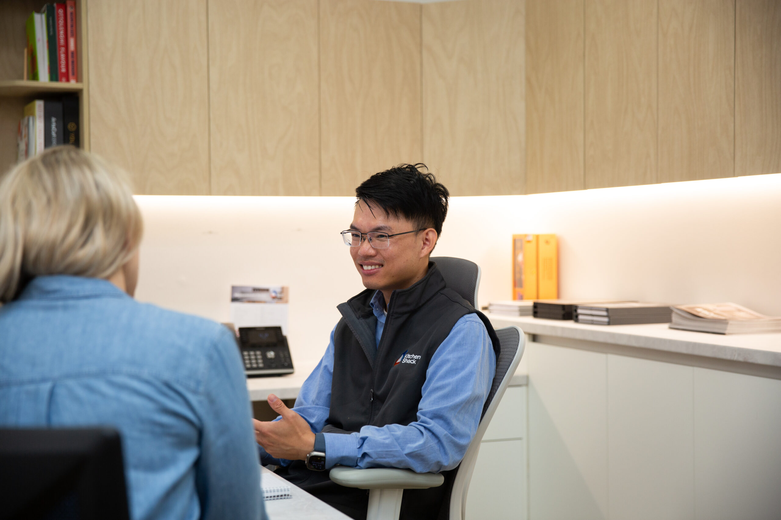Kitchen Designer dressed in a blue shirt and vest in a kitchen showroom designing a kitchen for a lady at a desk