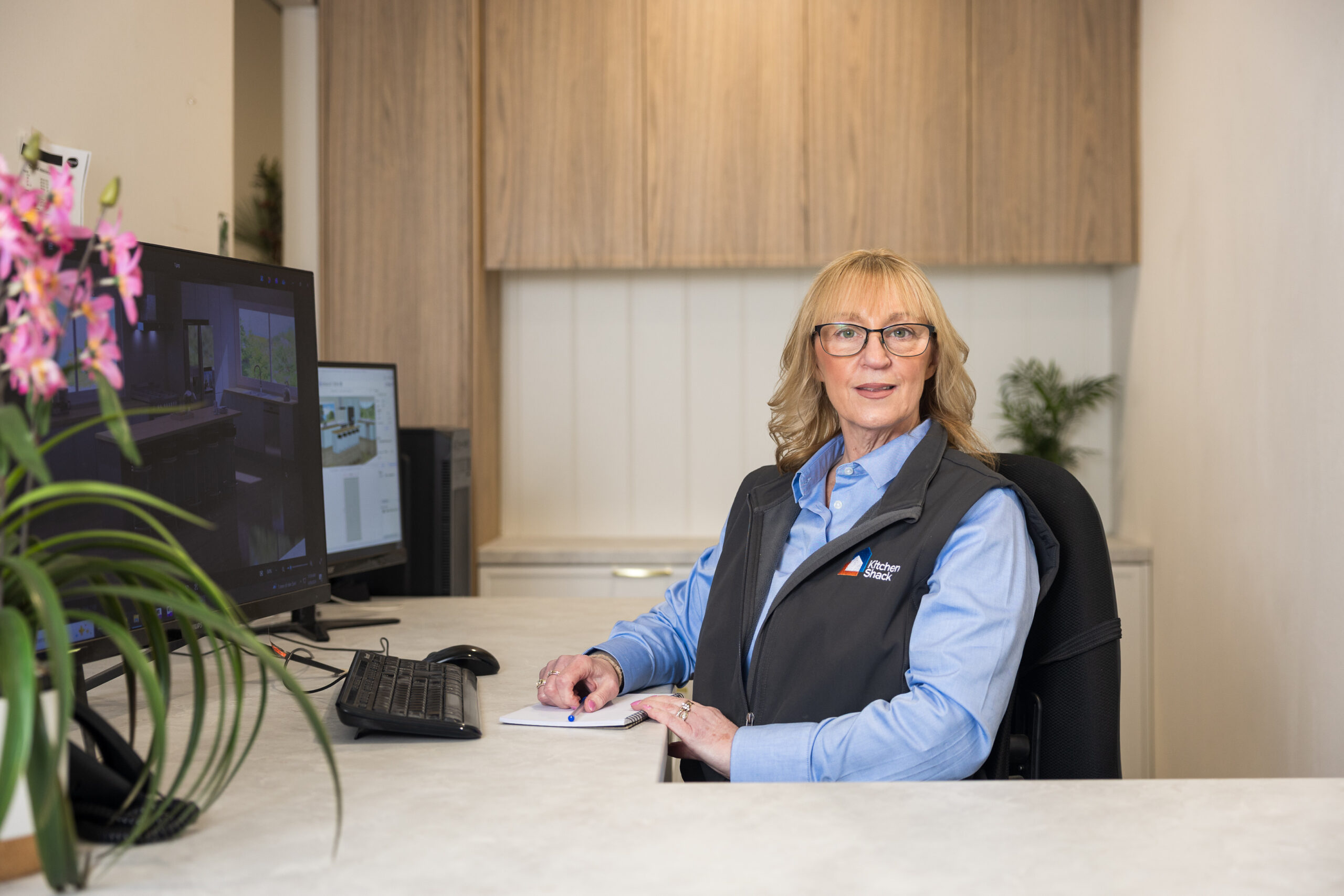 Lady in blue shirt and grey vest sitting at a desk designing a kitchen at a kitchen showroom with oak kitchen cabinetry behind