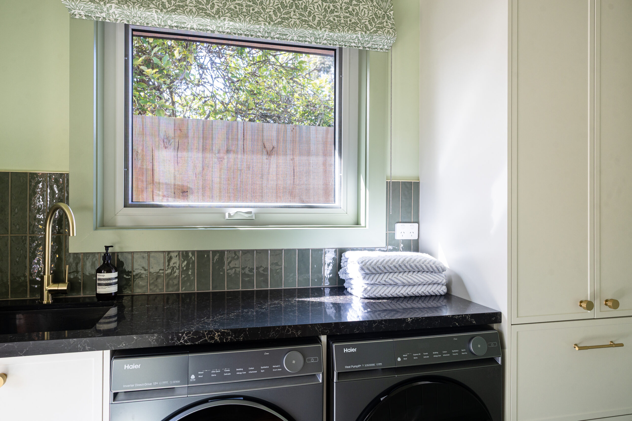 Galley shaped laundry with Hampton parchment cabinetry and brass handles with black washer dryer side by side and black benchtops and sink