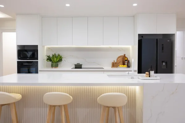 Open plan kitchen in white kitchen cabinetry and large staone island bench with waterfall ends, vase of flowers and chopping board with oils and lemons on benchtop