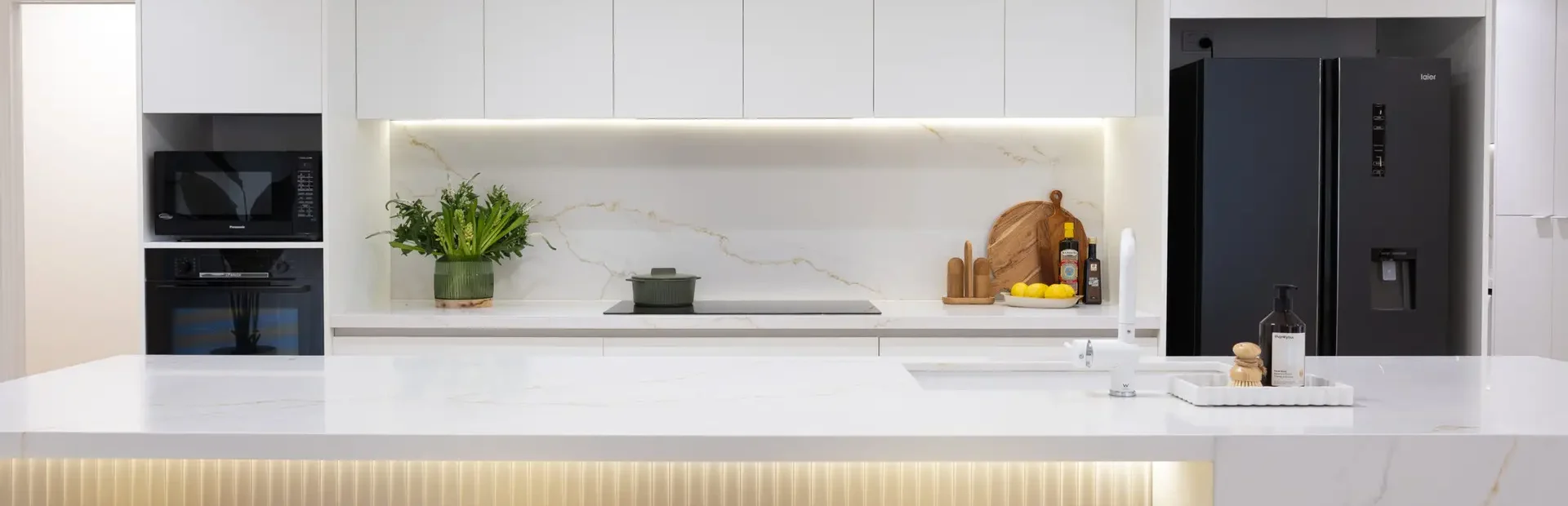 Open plan kitchen in white kitchen cabinetry and large staone island bench with waterfall ends, vase of flowers and chopping board with oils and lemons on benchtop
