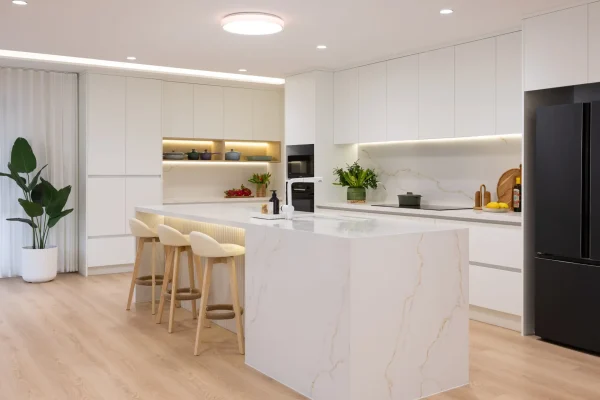 Large open plan kitchen featuring white cabinetry and standalone stone island bench with black oven and microwave and vase of flowers and pots decorating benchtops and open shelf