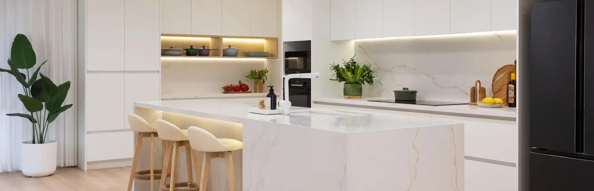 Large open plan kitchen featuring white cabinetry and standalone stone island bench with black oven and microwave and vase of flowers and pots decorating benchtops and open shelf