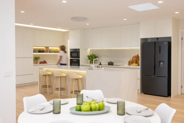 Female walking through a large open plan kitchen galley style with white cabinetry and stone benchtops displaying a round table and chairs in nearby dining room and three bar stools at breakfast bar