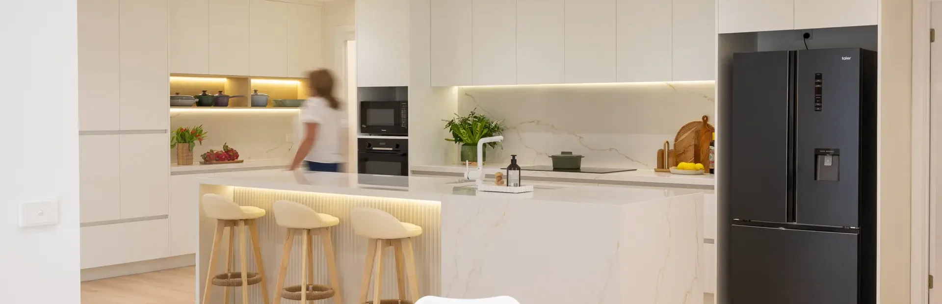 Female walking through a large open plan kitchen galley style with white cabinetry and stone benchtops displaying a round table and chairs in nearby dining room and three bar stools at breakfast bar