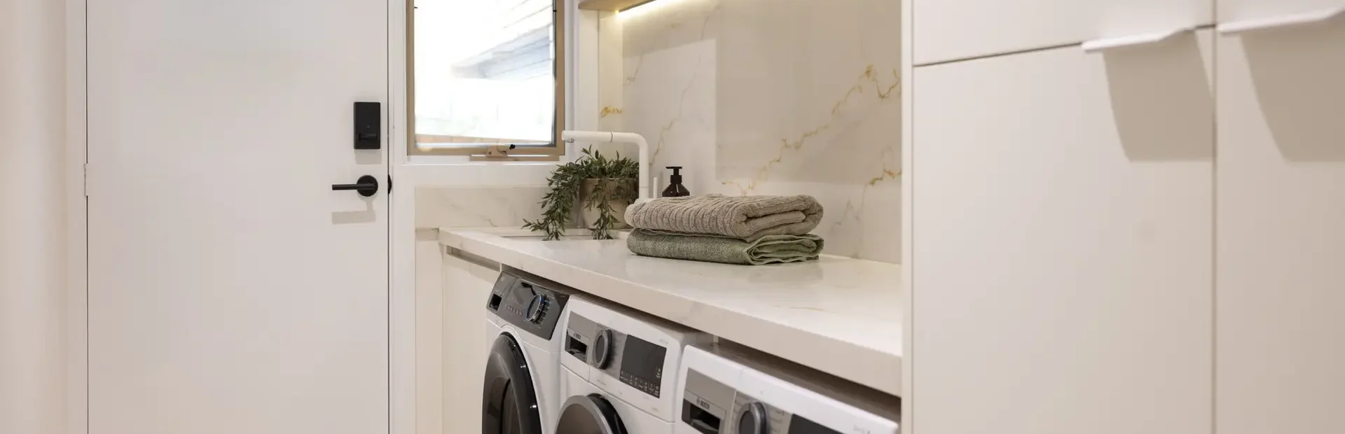 Galley laundry with white cabinetry and side by side washer dryers with green towels and hand soap on stone benchtop
