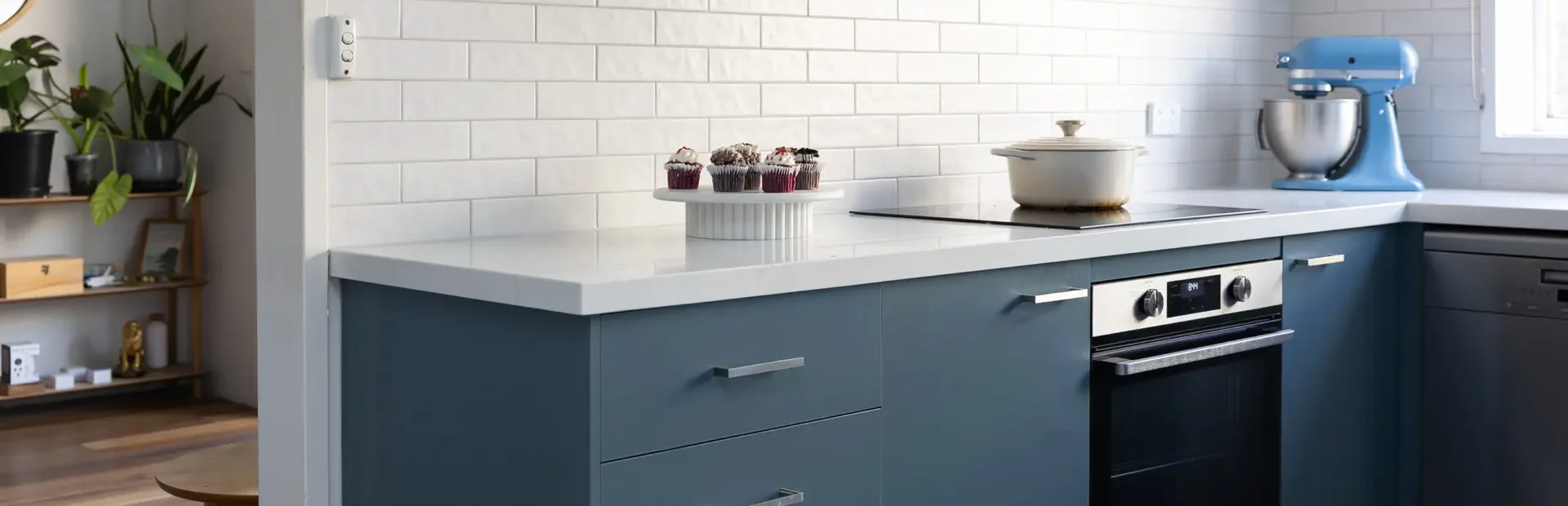 Blue kitchen cabinetry with chrome handles and black underbench oven with cream pot on cooktop and cakes on a cake stand