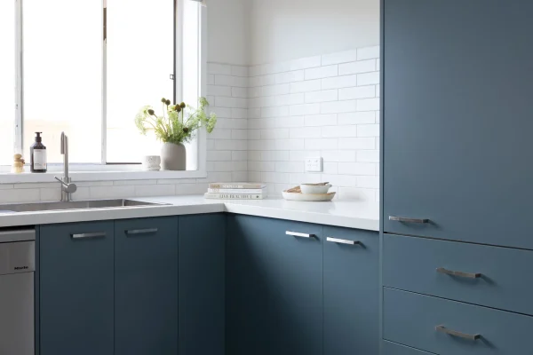 Corner blue cabinetry in kitchen with white stone benchtops and flowers in vase on window ledge