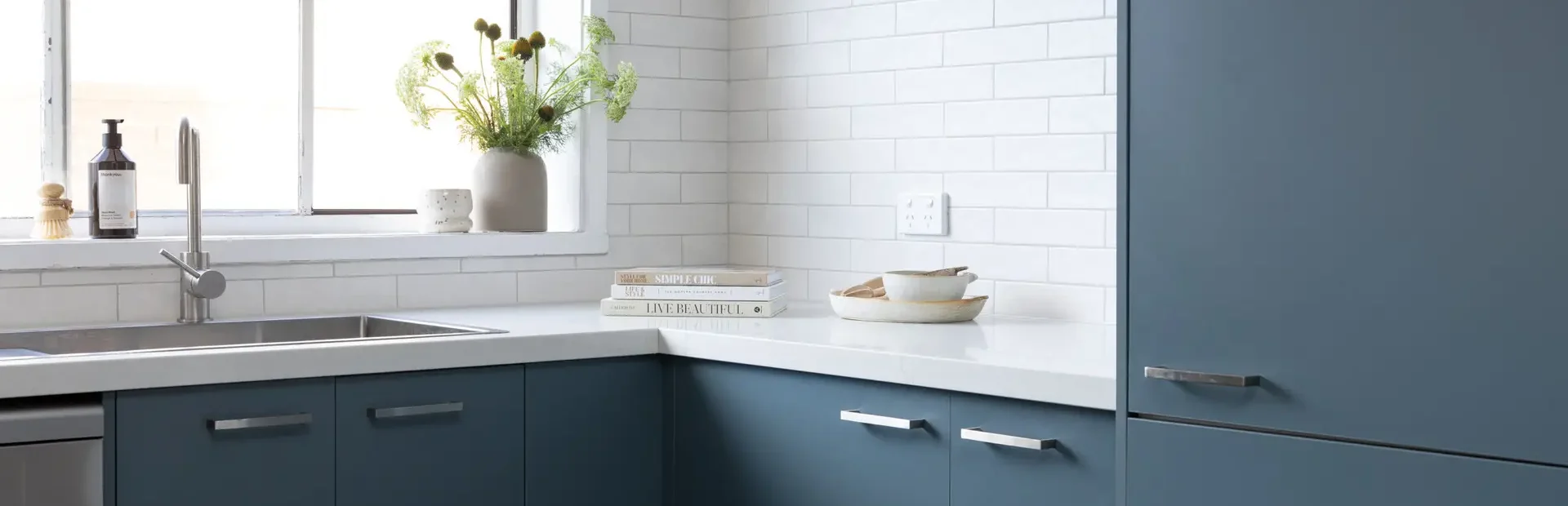 Corner blue cabinetry in kitchen with white stone benchtops and flowers in vase on window ledge