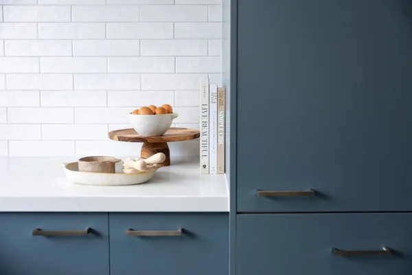 Blue kitchen cabinetry with chrome handles, white benchtops with styling books, eggs and pottery on bench