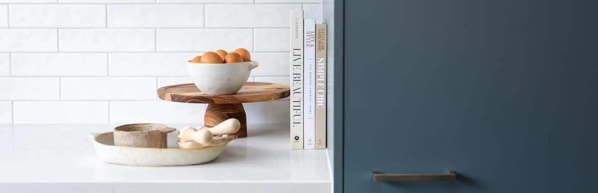 Blue kitchen cabinetry with chrome handles, white benchtops with styling books, eggs and pottery on bench