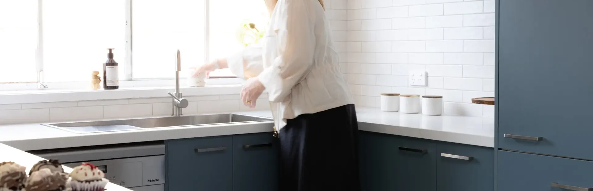 Female organising items in a blue u shape kitchen at the sink with open window