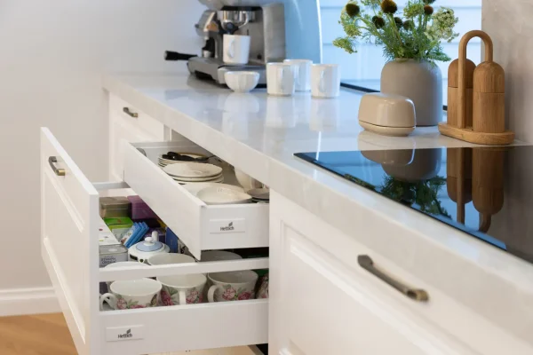 White carrington style kitchen cabinetry featuring drawers with inner concealed drawer with teas and cups, coffee machine on stone benchtop with coffee cups and country road vase of flowers with butter dish