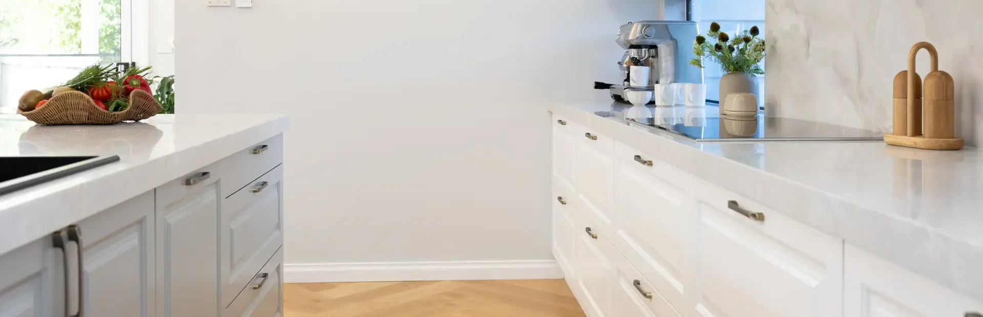 Galley style kitchen featuring white and grey kitchen cabinetry in carrington style with chrome handles and coffee machine with cups on benchtops as well as vase of flowers and styling items