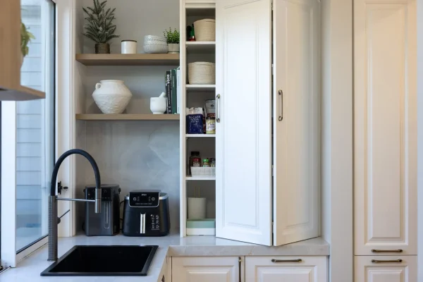 White kitchen cabinetry in appliance cupboard with oak shelving featuring vases and styling items with black sink and tap