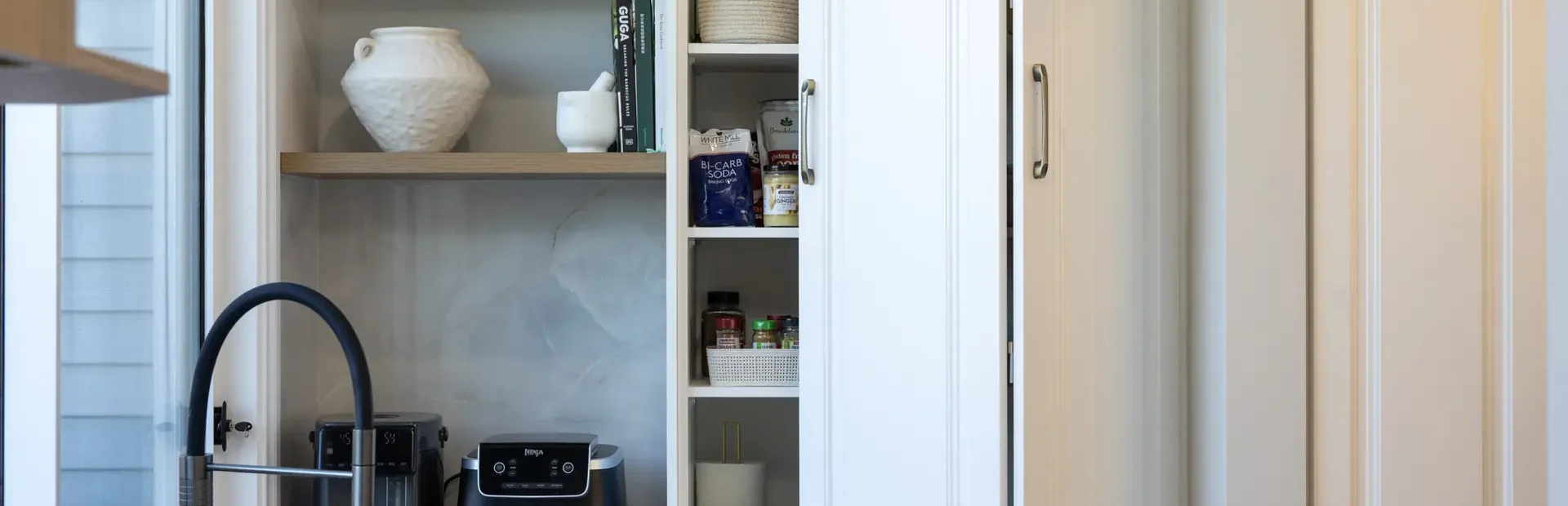 White kitchen cabinetry in appliance cupboard with oak shelving featuring vases and styling items with black sink and tap