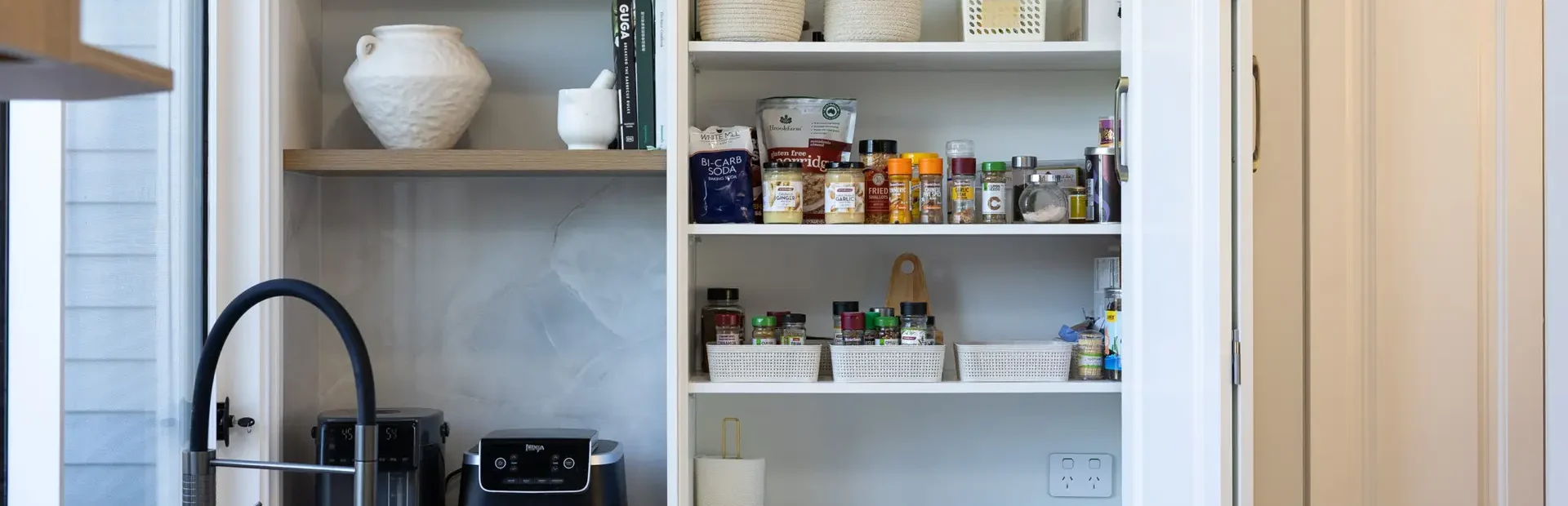 Butlers pantry featuring appliance cupboard in white shaker style cabinetry with pantry items, herbs and spices next to corner shelves in oak and black airfryer next to black sink and gooseneck tap