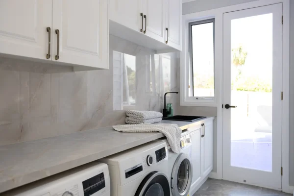 Galley style laundry featuring white carrington cabinetry underbench and overhead cabinetry with stone benchtops with door leading to outdoors, pile of towels on benchtops with washer dryer combinations under bench
