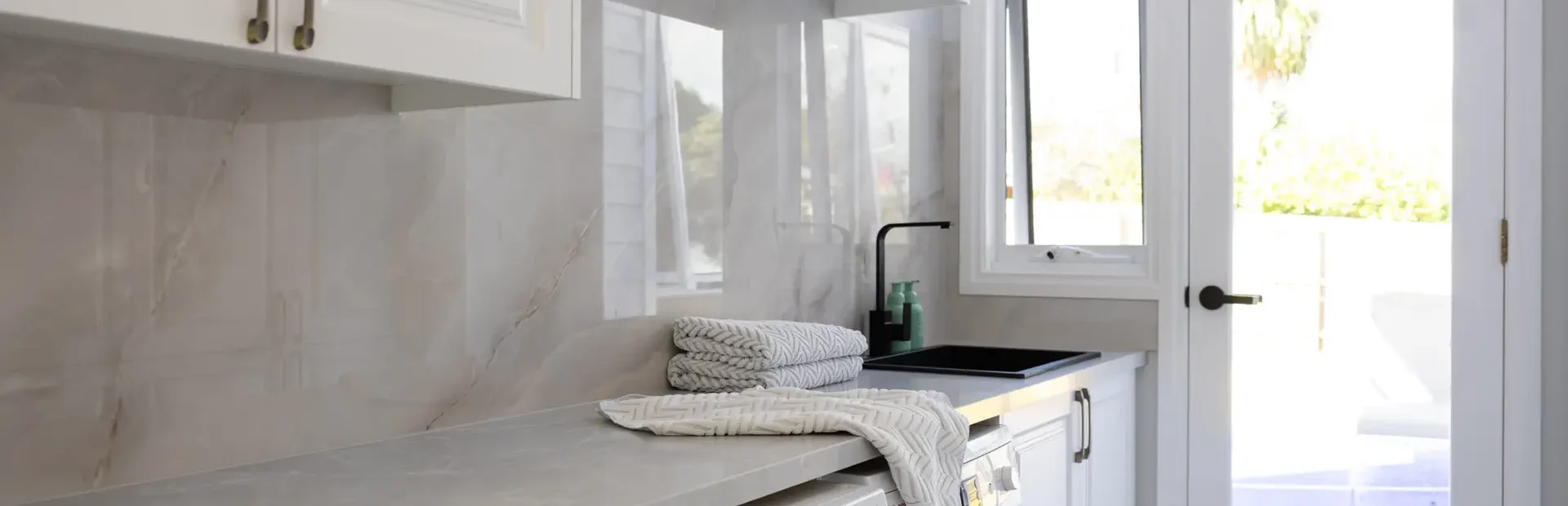 Galley style laundry featuring white carrington cabinetry underbench and overhead cabinetry with stone benchtops with door leading to outdoors, pile of towels on benchtops with washer dryer combinations under bench