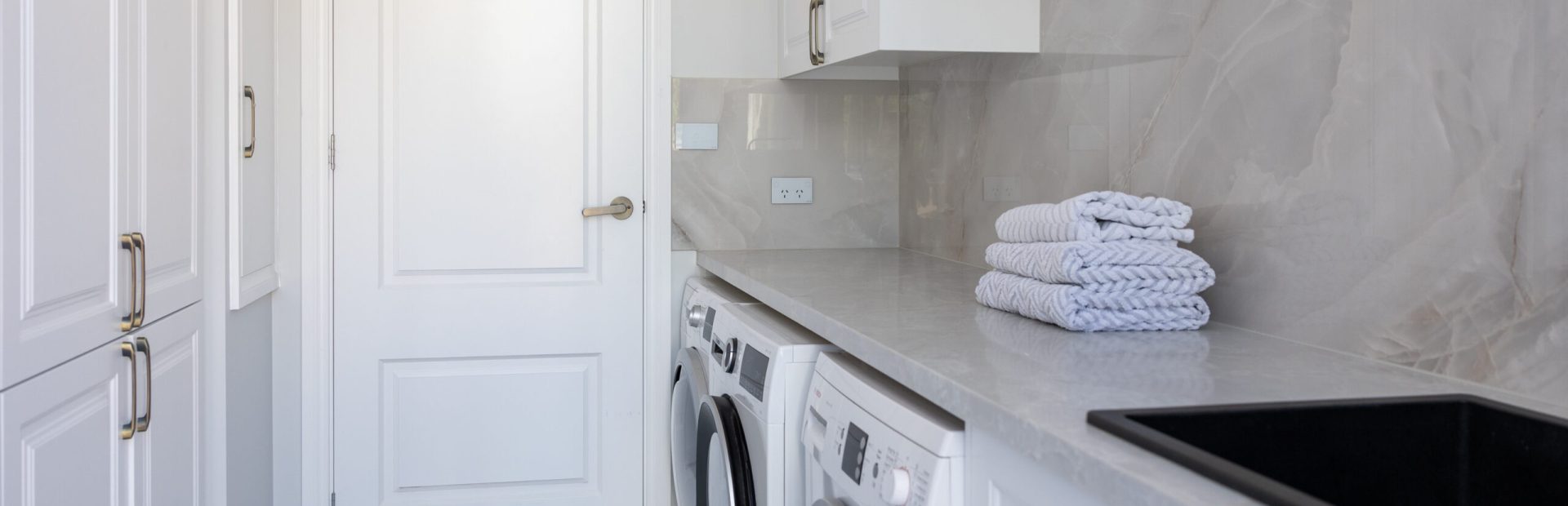 Laundry in galley style featuring white shaker style cabinetry in underbench and overheads on stone benchtops and splashbacks with washer dryer combination and black sink