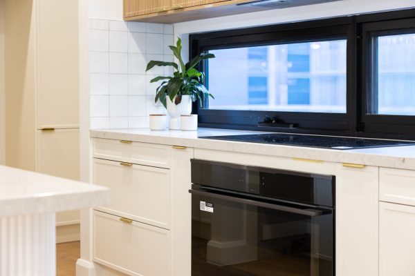 Hampton style kitchen drawers set of 3 with brass handles next to a pull out spice rack and black oven with black windows and plant on stone benchtop in white