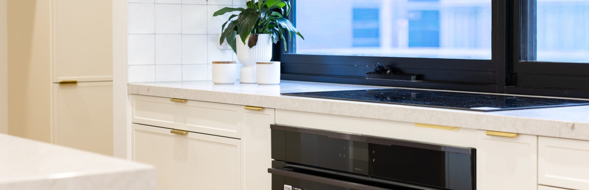 Hampton style kitchen drawers set of 3 with brass handles next to a pull out spice rack and black oven with black windows and plant on stone benchtop in white