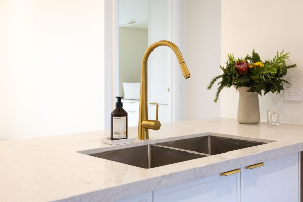 White kitchen cabinetry with metal sink and brass tapware with flowers and hand soap on stone benchtop